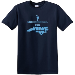 K&ouml;nnte beinhalten: Marineblaues T-Shirt mit hellblauer Grafik. Die Grafik zeigt einen Fu&szlig;abdruck, die Worte "UNCONDITIONAL FAN" und ein Basketball-Design &uuml;ber einer Umrisslinie des Bundesstaates North Carolina. Ein Gildan-Etikett ist sichtbar.