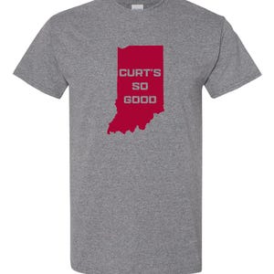 May include: A heather gray t-shirt featuring a red outline of the state of Indiana. The text "CURT'S SO GOOD" is printed in white within the state's borders. The shirt has short sleeves and a crew neck.