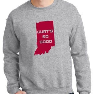 May include: A heather gray sweatshirt with a red graphic of the state of Indiana. The graphic includes the text "CURT'S SO GOOD" in white letters. The sweatshirt is a crew neck style.