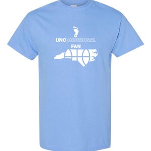 K&ouml;nnte beinhalten: Hellblaues T-Shirt mit wei&szlig;er Grafik. Die Grafik zeigt einen Fu&szlig;abdruck, die Worte "UNCONDITIONAL FAN" und eine stilisierte Abbildung des Bundesstaates North Carolina mit einem Basketball-Design. Das T-Shirt hat kurze &Auml;rmel und einen Rundhalsausschnitt.