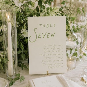 May include: A white rectangular table number card with green calligraphy reading Table Seven and a list of names including Olivia Gallego, Warner Stroupe, Charlotte Feig, William Fitzgerald, Estelle Darcy, Patrick Andrade, Cameron Mercer, Greta Evans.