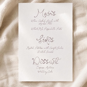 May include: A rectangular white menu card featuring dark grey cursive text. The headings are Mains, Sides, and Dessert. The listed items are Slow Cooked Lamb with Herbed Feta, Short Rib Pappardelle Pasta, Roasted Potatoes with Confit Garlic, Dutch Carrots, Espresso Martinis, and Wedding Cake.