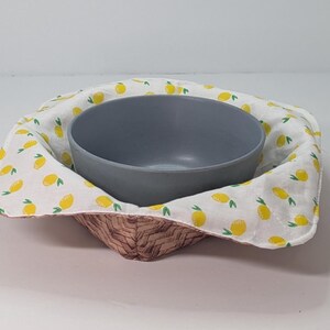 May include: A gray bowl nested inside a woven basket lined with a white fabric featuring a pattern of yellow lemons and green leaves. The basket has a brown and tan woven design.