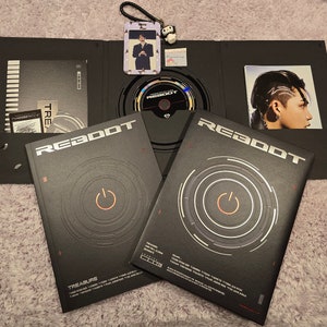 Puede incluir: Un conjunto de &aacute;lbum negro con la palabra "REBOOT" en blanco y naranja. La portada del &aacute;lbum presenta un dise&ntilde;o circular con un s&iacute;mbolo de bot&oacute;n de encendido. Incluye un CD, tarjetas fotogr&aacute;ficas y un peque&ntilde;o llavero.