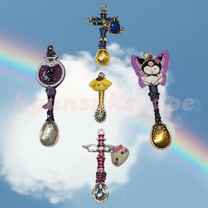 Puede incluir: Una colección de cucharas decorativas con varios amuletos. Los amuletos incluyen un gato negro en una botella de poción, un planeta, un personaje amarillo, un personaje de mariposa y una cabeza de Hello Kitty. Cada cuchara tiene un mango decorativo y un cuenco.
