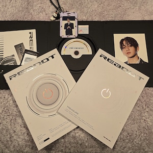 Puede incluir: Un conjunto de &aacute;lbumes en blanco y negro con la palabra "REBOOT" en letras plateadas en negrita. El conjunto incluye un CD, tarjetas fotogr&aacute;ficas y un cord&oacute;n con un amuleto. Las portadas del &aacute;lbum presentan un dise&ntilde;o circular con un s&iacute;mbolo de encendido.