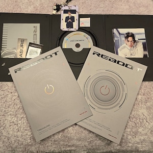 Puede incluir: Un conjunto de &aacute;lbum negro abierto que presenta un CD, tarjetas fotogr&aacute;ficas y un folleto. La portada del &aacute;lbum es de color gris claro con la palabra "REBOOT" en plata y un dise&ntilde;o circular con un s&iacute;mbolo de encendido. El &aacute;lbum est&aacute; etiquetado como "TREASURE".