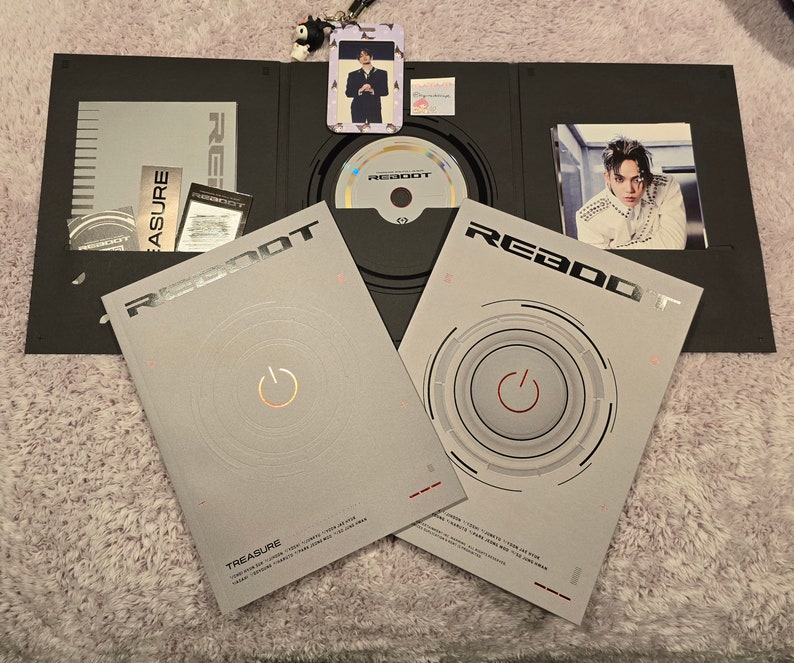 Puede incluir: Un conjunto de &aacute;lbum negro abierto que presenta un CD, tarjetas fotogr&aacute;ficas y un folleto. La portada del &aacute;lbum es de color gris claro con la palabra "REBOOT" en plata y un dise&ntilde;o circular con un s&iacute;mbolo de encendido. El &aacute;lbum est&aacute; etiquetado como "TREASURE".