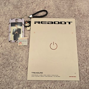 Puede incluir: Una portada de &aacute;lbum blanca con la palabra "REBOOT" en negro, un s&iacute;mbolo de encendido rojo y la palabra "TREASURE" en la parte inferior. Tambi&eacute;n hay una tarjeta fotogr&aacute;fica, un llavero con una correa trenzada negra y una peque&ntilde;a pegatina.