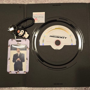 Puede incluir: Un &aacute;lbum negro con un CD y una tarjeta fotogr&aacute;fica. El CD est&aacute; etiquetado como "REBOOT". Tambi&eacute;n se ve un llavero con un personaje de dibujos animados en blanco y negro y un tarjetero con borde morado.