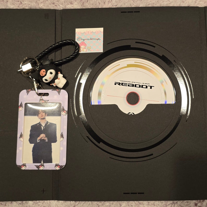 Puede incluir: Un &aacute;lbum negro con un CD y una tarjeta fotogr&aacute;fica. El CD est&aacute; etiquetado como "REBOOT". Tambi&eacute;n se ve un llavero con un personaje de dibujos animados en blanco y negro y un tarjetero con borde morado.