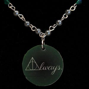 May include: A silver chain necklace with a dark green pendant. The pendant is round with the word "Always" written in white script, and a triangle symbol. The necklace has gray beads and green and blue crystals.