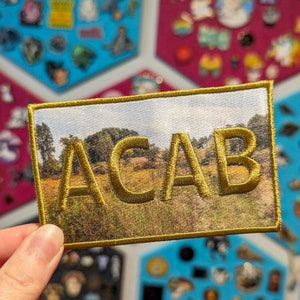 Peut inclure: Un écusson brodé or avec l'acronyme "ACAB" sur un fond de photo de paysage flou. L'écusson est entouré d'une bordure dorée.