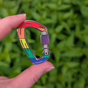 Peut inclure: Épingle en forme de mousqueton aux couleurs de l'arc-en-ciel. Le design comprend du rouge, de l'orange, du jaune, du vert et du bleu. Le mécanisme de verrouillage est violet. L'épingle est tenue par les doigts d'une personne.