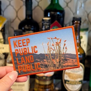 May include: Rectangular patch with an orange border and the words "KEEP PUBLIC LAND PUBLIC" in orange block letters. The patch features a desert landscape with orange plants and a blue sky.
