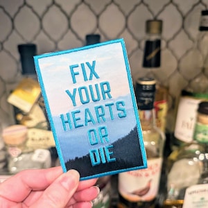 Op de afbeelding: Blauw en zwart geborduurd patch met de tekst "Fix Your Hearts Or Die" tegen een blauw en wit berglandschap.