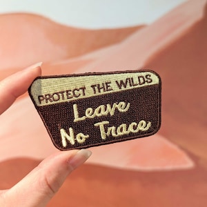 Pode incluir: Etiqueta bordada marrom e creme com o texto "Protect the Wilds Leave No Trace".