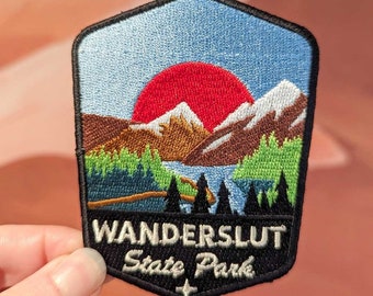 May include: Embroidered patch with a sunset scene over mountains and a lake. The patch is black with white text that reads "Wanderslut State Park".