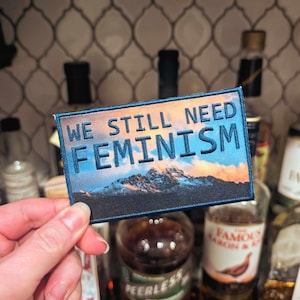 Puede incluir: Un parche bordado azul y blanco con un fondo de cordillera y el texto "WE STILL NEED FEMINISM".