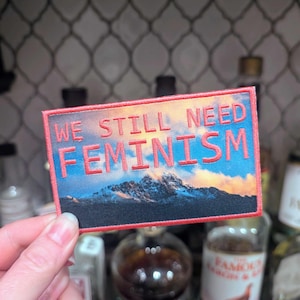 Op de afbeelding: Een rood omrande patch met de tekst "WE STILL NEED FEMINISM" in witte letters. De achtergrond van de patch is een foto van een bergketen met een bewolkte lucht.