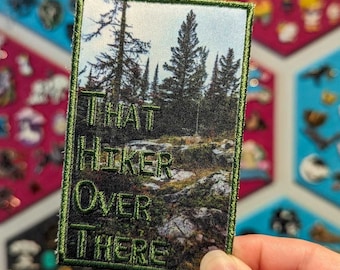 Puede incluir: Parche bordado verde con una foto de una escena de bosque y el texto "THAT HIKER OVER THERE" en blanco.