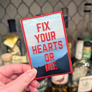 Könnte beinhalten: Roter und blauer gestickter Aufnäher mit dem Text "Fix Your Hearts Or Die" vor dem Hintergrund eines blauen Himmels und grüner Bäume.