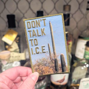 Può includere: Toppa ricamata con la scritta "DON'T TALK TO I.C.E." su uno sfondo di cielo blu con cactus del deserto. La toppa ha un bordo dorato ed è di forma rettangolare.