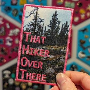 May include: Embroidered patch with a landscape scene of a forest and the text "THAT HIKER OVER THERE" in pink. The patch has a pink border and is rectangular in shape. The background is a mix of green, brown, and blue.