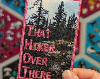 Puede incluir: Parche bordado con un paisaje forestal y el texto rosa "THAT HIKER OVER THERE". El parche rectangular tiene un borde rosa. El fondo muestra una mezcla de verde, marrón y azul, que representa una escena natural.