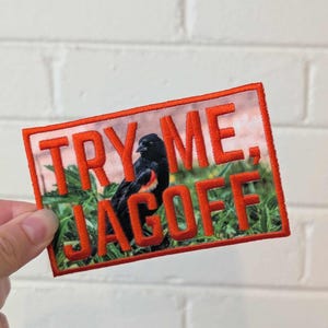 May include: Rectangular patch with a red border and text that reads "TRY ME, JAGOFF" in red. The patch features a bird with black feathers and a red shoulder, perched in green foliage.