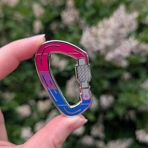 Puede incluir: Pin de mosquetón metálico con una combinación de colores rosa, morado y azul. El pin se sostiene en una mano sobre un fondo borroso verde y blanco. El mosquetón tiene un mecanismo de bloqueo plateado.