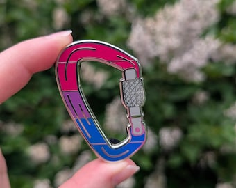 May include: A metal carabiner pin with a pink, purple, and blue color scheme. The pin is held in a hand against a blurred green and white background. The carabiner has a silver locking mechanism.
