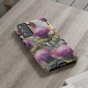 Puede incluir: Una funda de teléfono con un diseño floral con flores de cardo púrpura sobre un fondo blanco.