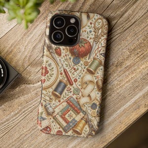 May include: A smartphone case featuring a detailed vintage sewing pattern with illustrations of a red tomato pincushion, wooden thread spools, metal scissors, buttons, a thimble, and a quilted fabric square on a cream-colored background.