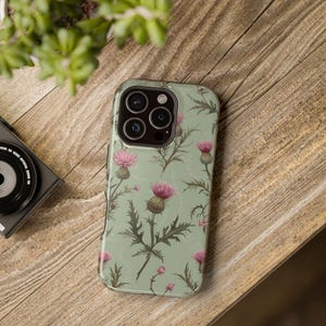 Puede incluir: Una funda de teléfono con un diseño floral de cardo. La funda presenta un fondo verde claro con cardos rosas y hojas verdes. La funda del teléfono está sobre una superficie de madera.
