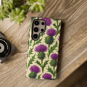 May include: A phone case featuring a repeating pattern of purple thistle flowers with green leaves on a cream background. The case is shown on a wooden surface, with a camera and plant in the background.