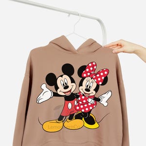 Może przedstawiać: Beżowa bluza z kapturem z grafiką Myszki Miki i Myszki Minnie. Miki ma na sobie czerwone szorty, a Minnie czerwoną sukienkę w kropki i kokardę. Bluza ma napis "Lemon & Co" na dole.