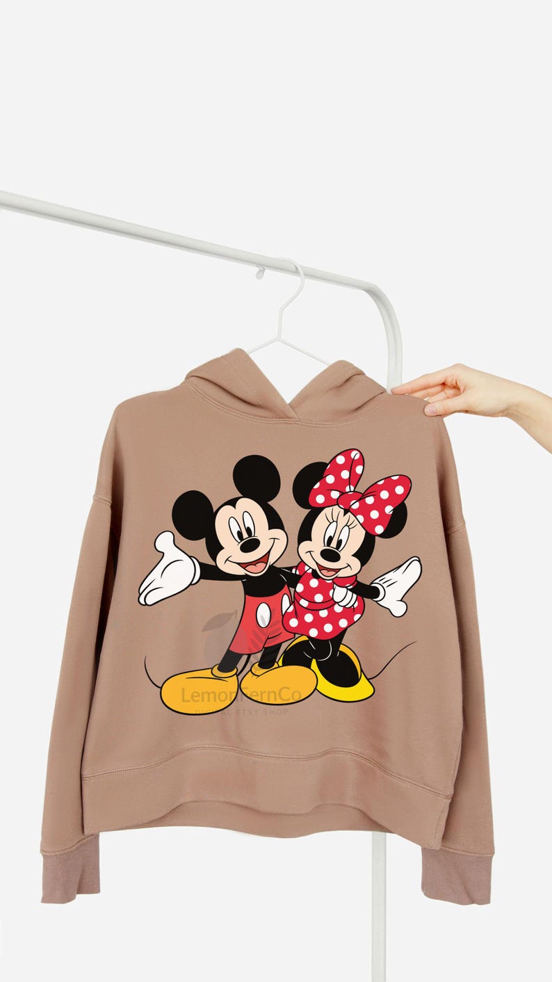 Może przedstawiać: Beżowa bluza z kapturem z grafiką Myszki Miki i Myszki Minnie. Miki ma na sobie czerwone szorty, a Minnie czerwoną sukienkę w kropki i kokardę. Bluza ma napis "Lemon & Co" na dole.