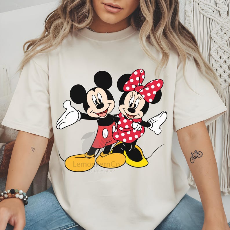Może przedstawiać: Kremowa koszulka z Myszka Miki i Myszka Minnie. Miki ma na sobie czerwone szorty i ż&oacute;łte buty, a Minnie czerwoną sukienkę w kropki i kokardę. Koszulka ma napis "LemoncornCo" na dole.