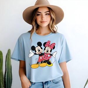 Może przedstawiać: Jasnoniebieska koszulka z grafiką Myszki Miki i Myszki Minnie. Miki ma na sobie czerwone szorty, a Minnie czerwoną kokardę i sukienkę w kropki. Koszulka ma napis "Lemon Tree Co" na dole.