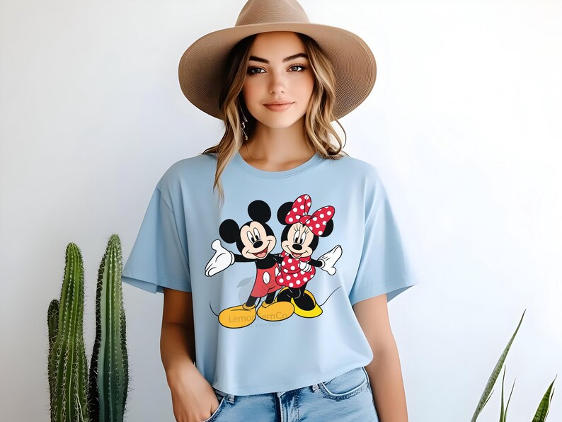 Może przedstawiać: Jasnoniebieska koszulka z grafiką Myszki Miki i Myszki Minnie. Miki ma na sobie czerwone szorty, a Minnie czerwoną kokardę i sukienkę w kropki. Koszulka ma napis "Lemon Tree Co" na dole.