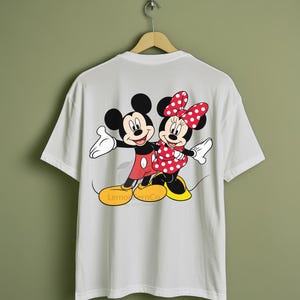 Może przedstawiać: Biała koszulka z kolorową grafiką Myszki Miki i Myszki Minnie w uścisku. Miki ma na sobie czerwone spodenki, a Minnie czerwoną sukienkę w kropki. Koszulka wisi na drewnianym wieszaku.