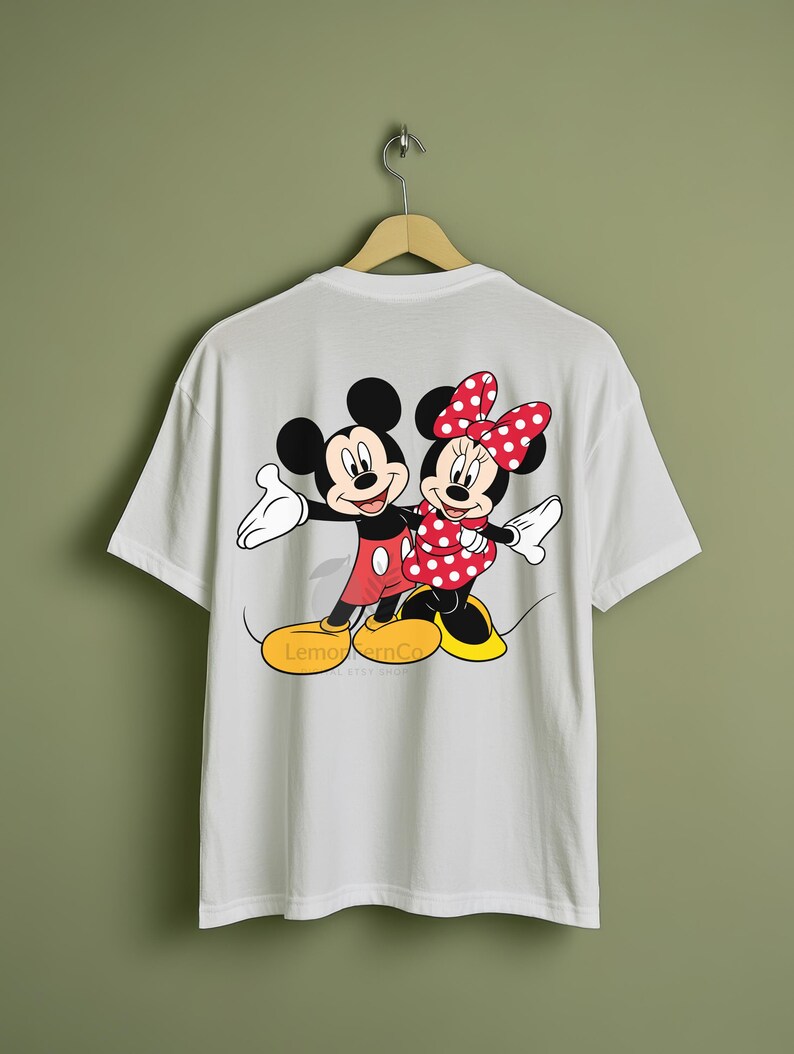 Może przedstawiać: Biała koszulka z kolorową grafiką Myszki Miki i Myszki Minnie w uścisku. Miki ma na sobie czerwone spodenki, a Minnie czerwoną sukienkę w kropki. Koszulka wisi na drewnianym wieszaku.