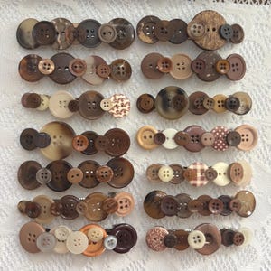 May include: A collection of twelve hair clips, each featuring a unique arrangement of assorted brown, tan, and cream-coloured buttons. The buttons vary in size, texture, and pattern, including solid, speckled, and checkered designs, securely attached.