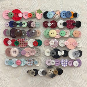 Pode incluir: Uma coleção de onze ganchos de cabelo decorados com vários botões coloridos de formas, tamanhos e padrões variados. Os botões incluem designs redondos, em forma de coração e com temas de frutas em tons de vermelho, rosa, azul, verde, roxo e preto.