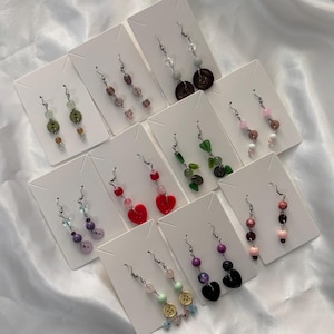 May include: A collection of handmade earrings displayed on white cards. The earrings feature various designs, including beads, buttons, and heart-shaped accents in a range of colors like red, green, purple, and pink. Each pair hangs from a silver-colored hook.