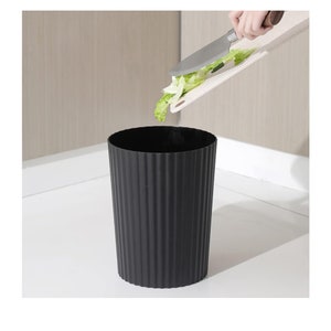 May include: A black, ribbed, cylindrical bin with a white chopping board and green lettuce being placed inside.