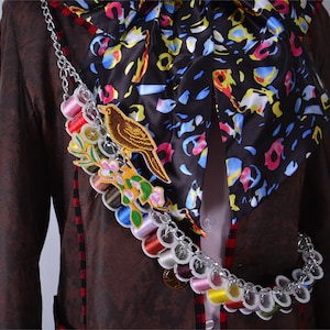 May include: A brown jacket with a colorful patterned scarf and a chain with spools of thread. The chain has a small bird and flower embroidery.