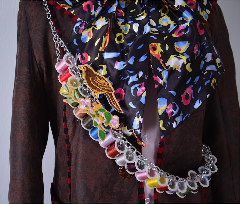 May include: A brown jacket with a colorful patterned scarf and a chain with spools of thread. The chain has a small bird and flower embroidery.