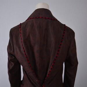 May include: Brown suede jacket with red and black trim. The jacket has a collar and long sleeves.